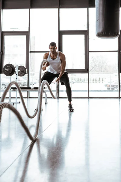 Selective focus of handsome sportsman exercising with battle ropes in modern gym — Stock Photo