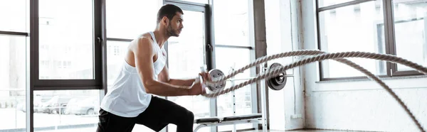 Panoramic shot of strong sportsman exercising with battle ropes in modern gym — Stock Photo