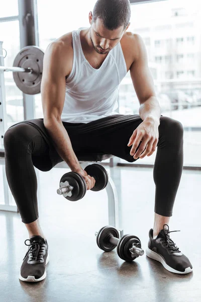 Bell'uomo sportivo che si allena con i manubri mentre è seduto in palestra — Foto stock