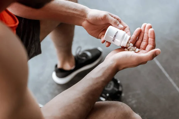 Обрезанный вид спортсмена, держащего бутылку с пробиотическими надписями и таблетками в спортзале — стоковое фото