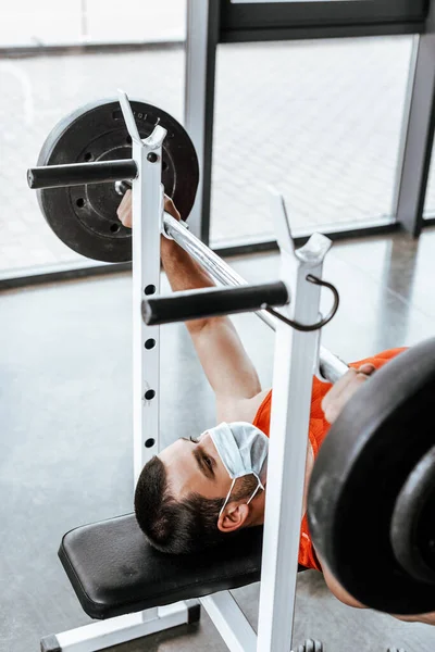 Selective focus of athletic sportsman in medical mask exercising with barbell in gym — Stock Photo