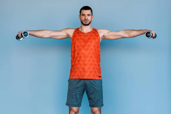 Deportista barbudo de pie con las manos extendidas y haciendo ejercicio con pesas en azul - foto de stock
