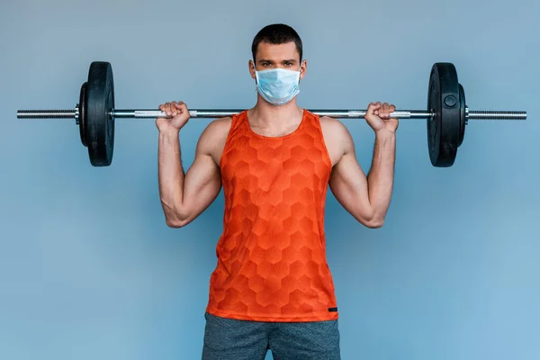 Deportista en máscara médica haciendo ejercicio con barra aislada en azul - foto de stock