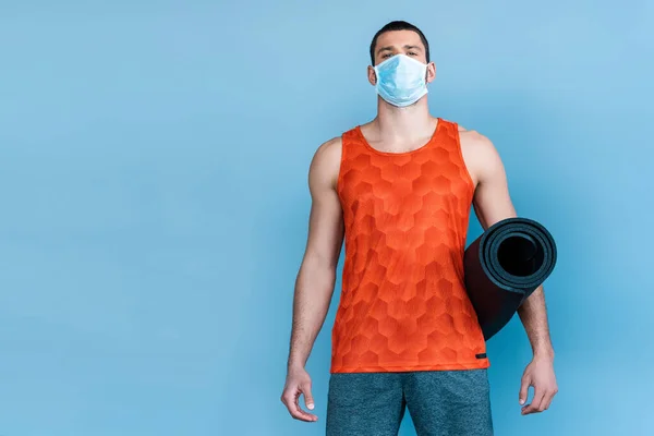 Sportif en masque médical debout avec tapis de fitness isolé sur bleu — Photo de stock