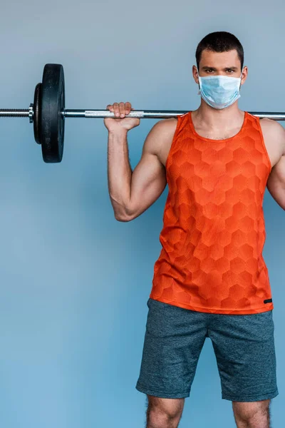 Deportista en máscara médica haciendo ejercicio con barra aislada en azul - foto de stock