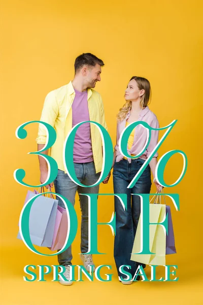 Красивая пара, держащая сумки с покупками и глядя друг на друга на желтый, 30 процентов скидка на весенние продажи иллюстрации — стоковое фото