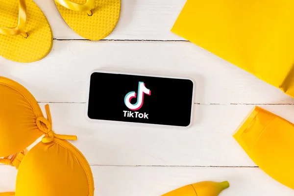 KYIV, UKRAINE - MARCH 25, 2020: Top view of frame from yellow flip flops with sunscreen, swimsuit and book near smartphone with TikTok app on white wooden planks — Stock Photo