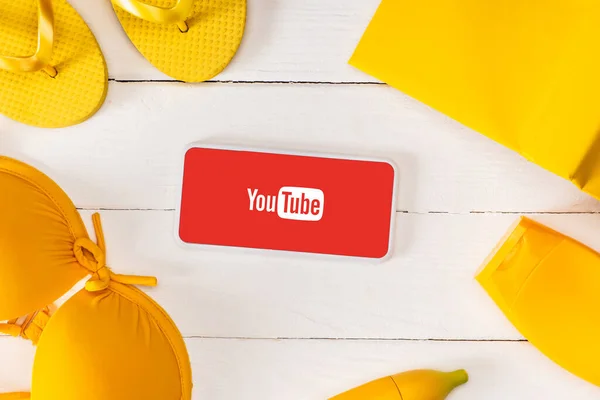 KYIV, UKRAINE - MARCH 25, 2020: Top view of frame from yellow flip flops with sunscreen, swimsuit and book near smartphone with youtube app on white wooden background — Stock Photo