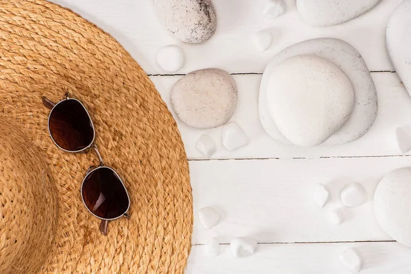 Vista superior de gafas de sol en sombrero de paja cerca de piedras marinas sobre fondo de madera blanca - foto de stock