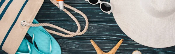 Panoramic shot of top view of sunglasses, flip flops near bag and starfish on dark wooden surface — Stock Photo
