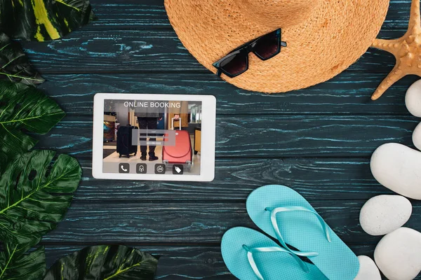 Draufsicht auf digitales Tablet mit Online-Buchungs-App in der Nähe von Flip Flops, Meeressteinen und Sonnenbrille auf dunkler Holzoberfläche — Stockfoto