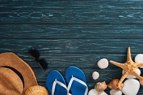 Vue de dessus des tongs, du chapeau de paille près des étoiles de mer et des cailloux sur des planches de bois foncé — Photo de stock