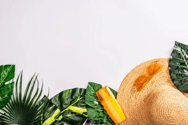 Vue du dessus du chapeau de paille près des lunettes de soleil et de la crème solaire sur les feuilles vertes sur la surface blanche — Photo de stock