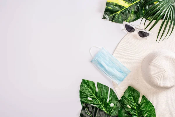 Vue du dessus du masque médical sur le chapeau de soleil et les lunettes de soleil avec des feuilles vertes sur la surface blanche — Photo de stock