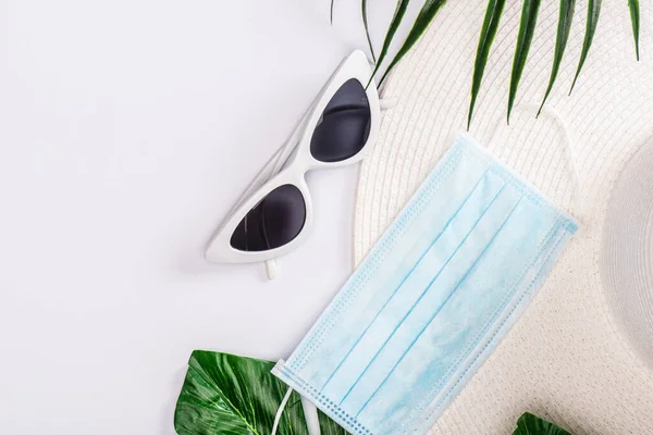 Draufsicht auf medizinische Maske mit Sonnenhut und Sonnenbrille in der Nähe grüner Blätter auf weißer Oberfläche — Stockfoto