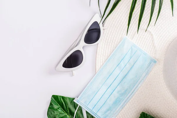 Top view of medical mask with sun hat and sunglasses near green leaves on white surface — Stock Photo