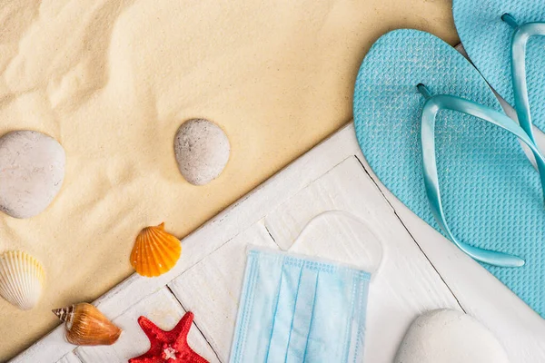 Draufsicht auf blaue Flip Flops in der Nähe medizinischer Maske und Muscheln auf weißen Holzplanken und Sand — Stockfoto