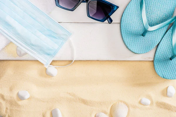 Vue du dessus du masque médical, des tongs et des lunettes de soleil sur des planches de bois blanc près de cailloux sur du sable — Photo de stock