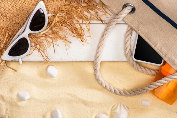 Vista superior de smartphone y protector solar en bolsa cerca de sombrero de paja y gafas de sol en tablones de madera blanca y guijarros en arena - foto de stock