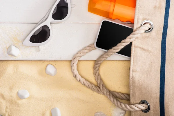 Vue du dessus du smartphone et de la crème solaire près du sac et des lunettes de soleil sur des planches de bois blanc et des pierres de mer sur du sable — Photo de stock