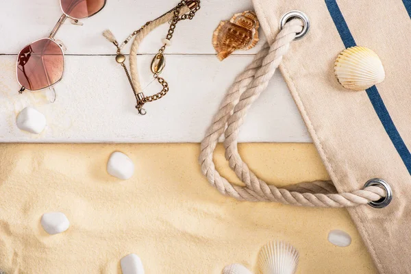 Vue de dessus des lunettes de soleil, bracelet et sac sur planches de bois blanc près de cailloux sur sable — Photo de stock