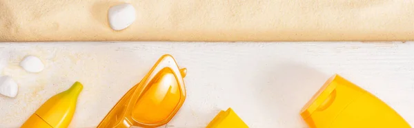 Панорамний знімок зверху жовтих сонцезахисних окулярів біля сонцезахисних кремів на білих дерев'яних дошках і гальки на піску — стокове фото