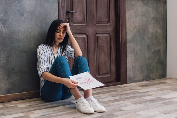 Расстроенная женщина смотрит на документ и сидит рядом со стеной на полу в комнате — стоковое фото