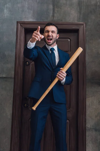 Angry collector with baseball bat pointing with finger and shouting near door in room — Stock Photo