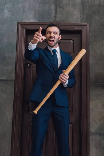 Coleccionista enojado con bate de béisbol apuntando con el dedo y gritando cerca de la puerta en la habitación - foto de stock