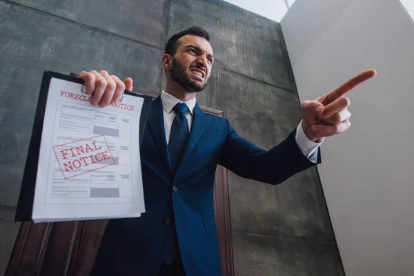 Low angle view of angry collector with documents with foreclosure and final notice lettering pointing with finger in room — Stock Photo