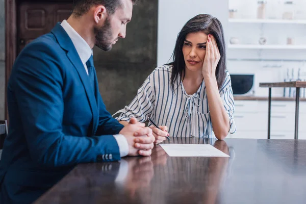 Mujer molesta sosteniendo pluma cerca de documento y mirando al coleccionista con las manos apretadas en la mesa en la habitación — Stock Photo