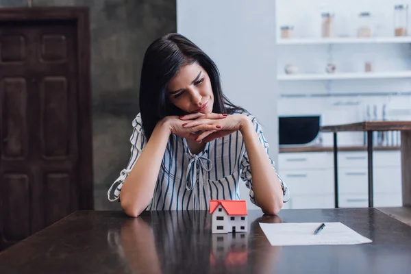 Upset and thoughtful woman with clenched hands at table with house model near document in room — Stock Photo