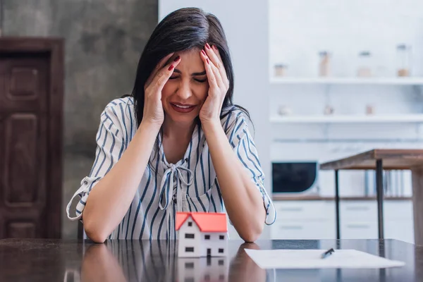 Empörte Frau weint an Tisch mit Hausmodell in Zimmernähe — Stockfoto