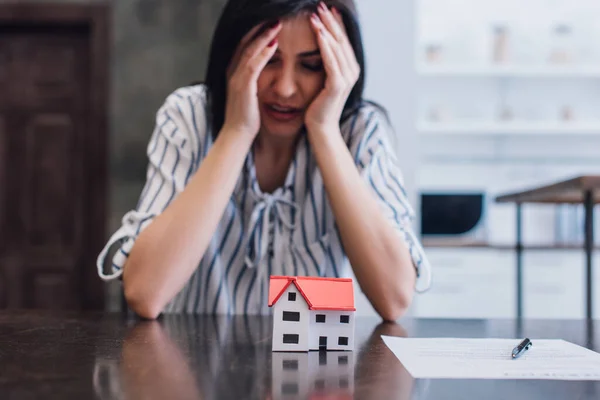 Femme bouleversée et stressée à table avec modèle de maison près du document avec stylo dans la chambre — Photo de stock