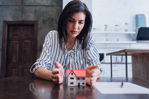Konzentrierte Frau legt Hände neben Hausmodell mit Papier und Stift auf Tisch im Zimmer — Stockfoto