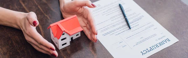 Ausgeschnittene Ansicht weiblicher Hände in der Nähe des Hausmodells, Papier mit Konkursschrift und Stift auf dem Tisch, Panoramaaufnahme — Stockfoto