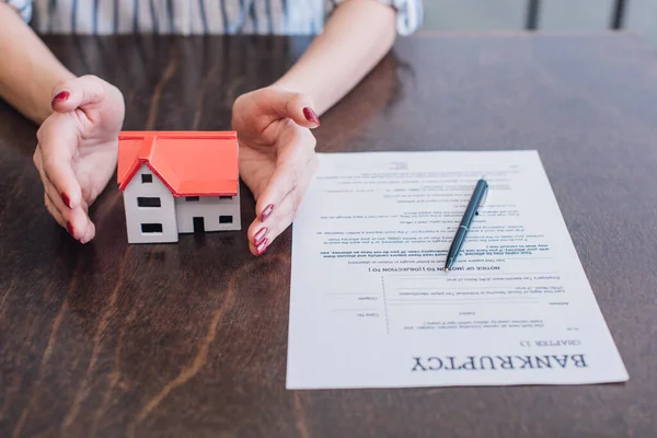 Vue partielle des mains féminines près du modèle de la maison, papier avec lettre de faillite et stylo sur la table — Stock Photo