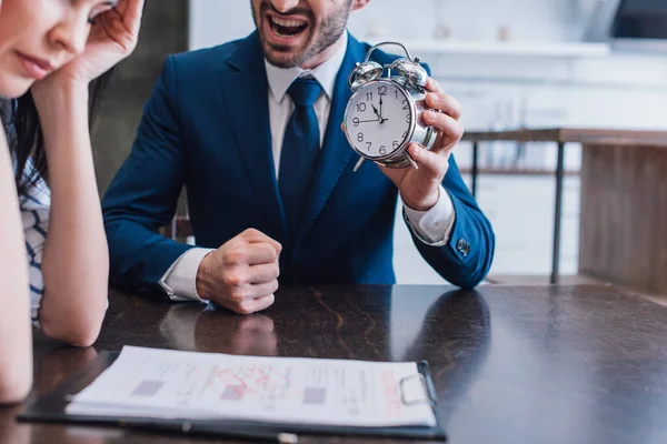 Cropped view of angry collector holding alarm clock near upset woman at table with documents in room — Stock Photo