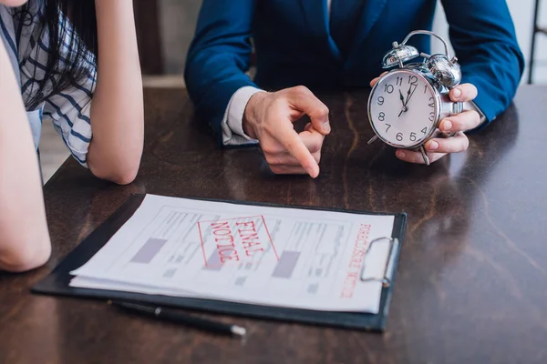 Cropped view of collector with alarm clock pointing at documents with foreclosure and final notice lettering near woman at table — Stock Photo