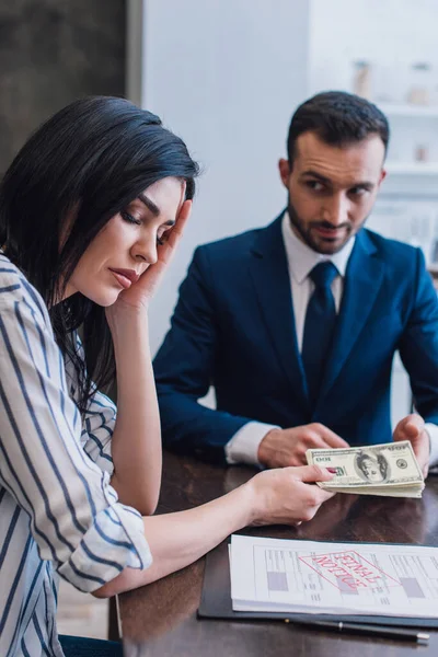 Selective focus of upset woman giving dollar banknotes to collector near documents and pen at table in room — Stock Photo