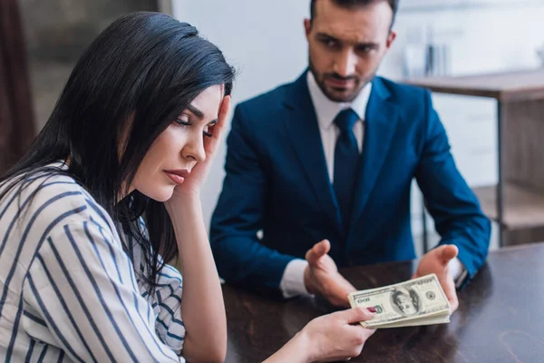 Selective focus of upset woman giving dollar banknotes to collector at table in room — Stock Photo