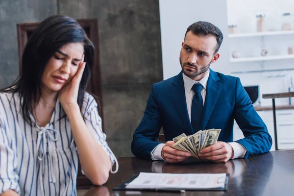 Mujer molesta llorando cerca de coleccionista con billetes de dólar en la mesa en la habitación - foto de stock