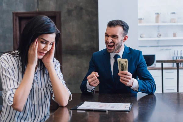 Coleccionista emocionado con billetes de dólar cerca de la mujer estresada en la mesa en la habitación - foto de stock