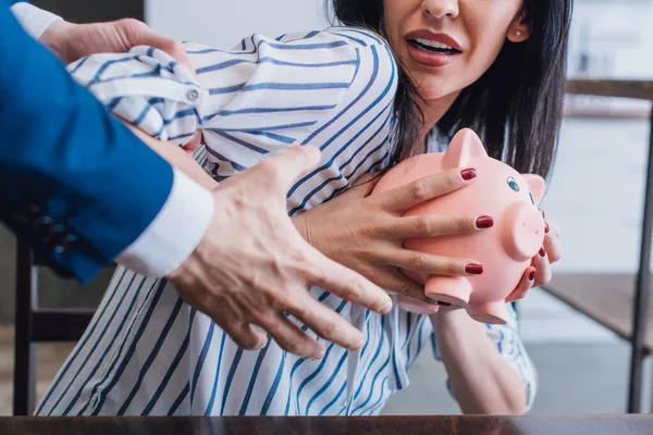 Vista recortada del coleccionista agarrando la mano femenina y la mujer asustada sosteniendo alcancía en la mesa en la habitación - foto de stock