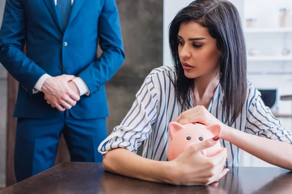 Mujer preocupada cubriendo alcancía con las manos en la mesa cerca de colector con las manos apretadas en la habitación - foto de stock