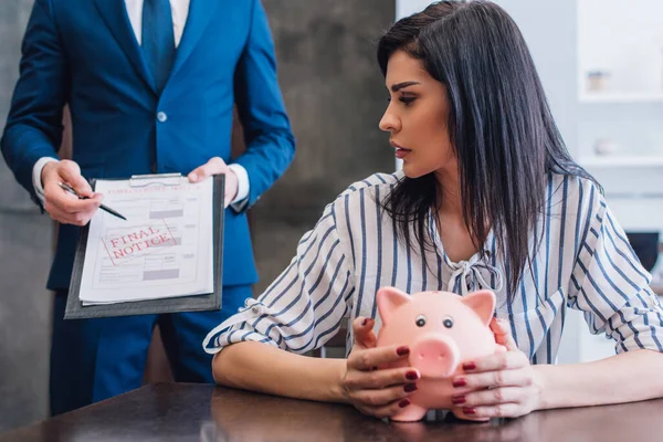 Femme avec tirelire à table près du collectionneur pointant avec stylo sur les documents avec dernier avis lettrage dans la chambre — Photo de stock