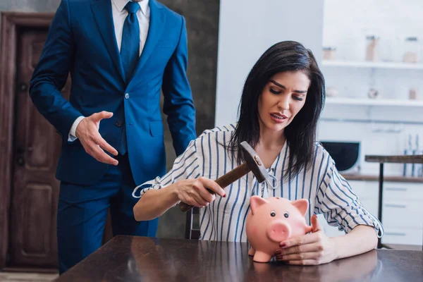 Besorgte Frau hält Hammer über Sparschwein am Tisch neben Sammler mit ausgestreckter Hand im Zimmer — Stockfoto