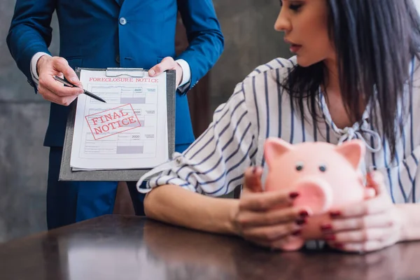 Cropped view of woman with piggy bank at table near collector pointing with pen at documents with final notice and foreclosure lettering in room — Stock Photo