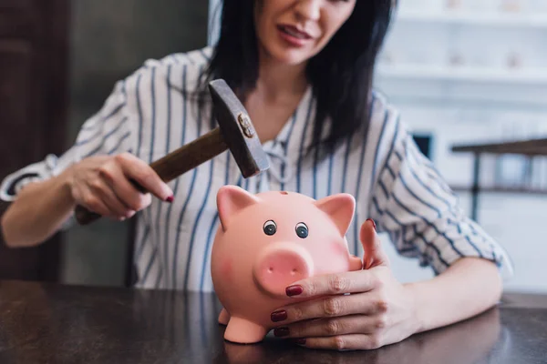 Cropped view of woman holding hammer above piggy bank at table — Stock Photo