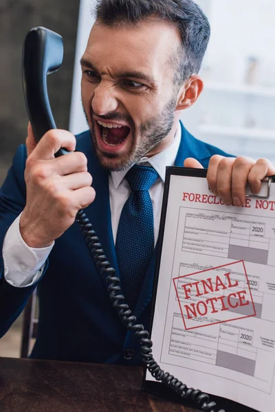 Aggressive collector shouting on handset and holding documents with foreclosure and final notice lettering at table in room — Stock Photo